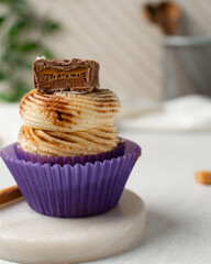 Close-up of a vanilla cupcake in a purple wrapper with buttercream frosting, cinnamon dust, and a caramel-filled chocolate, cupcake in a purple liner with a cinnamon frosting and caramel chocolate bar