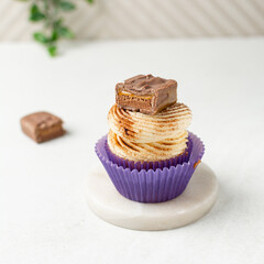 Close-up of a vanilla cupcake in a purple wrapper with buttercream frosting, cinnamon dust, and a caramel-filled chocolate, cupcake in a purple liner with a cinnamon frosting and caramel chocolate bar