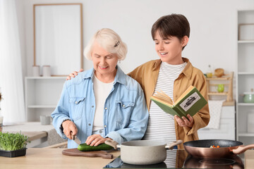 Senior woman cutting cucumber and her grandson with recipe book in kitchen