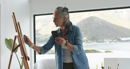 Woman referencing smartphone painting on canvas at wooden easel overlooking lake and mountain view