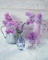 Bouquets of fresh lilac blossoms in rustic jugs and glass vases, spring flowers in the interior, wooden background.