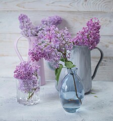 Bouquets of fresh lilac blossoms in rustic jugs and glass vases, spring flowers in the interior, wooden background.
