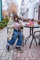 A young woman is reading a book in a street cafe, a bouquet of flowers is on the table, the woman is resting after shopping.