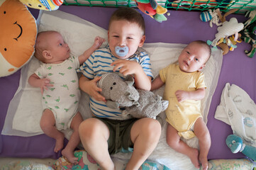 Three brothers enjoy their time on a mat. The older brother holds a stuffed toy while the twins lie beside him. They are surrounded by bright toys and a playful setting