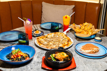 Gourmet cuisine assortment on restaurant table, featuring truffle pizza, pasta, fried seafood, and vibrant cocktails. Lively dining atmosphere, diverse culinary experience.