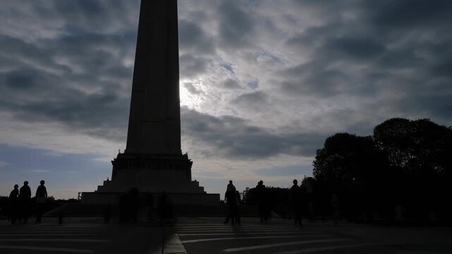 Dramatic low angle shot of a towering war memorial obelisk silhouetted against a cloudy, moody sky at dusk, with people moving solemnly around its base