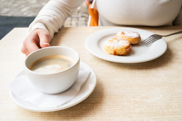Close-up of Caucasian woman enjoying warm drink with cottage cheese pancakes or syrniki at outdoor cafe table, girl enjoying her breakfast