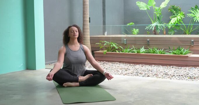 African adult woman in workout clothes breathing, lengthening spine on green mat at patio for calm