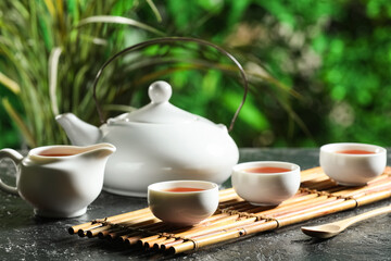 Bamboo mat with teapot, cups of tea and spoon for ceremony on black grunge table outdoors, closeup