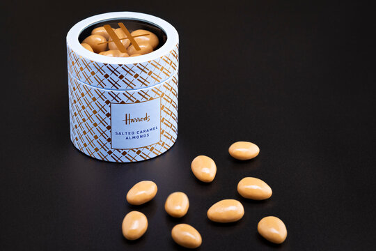 A premium tin of Harrods Salted Caramel Almonds accompanied by several glossy caramel-coated almonds The elegant patterned tin, featuring the iconic Harrods branding, reflects the luxury product..