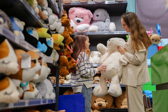 A young girl smiles as her mother holds a large teddy bear amidst shelves filled with various plush toys in a vibrant store
