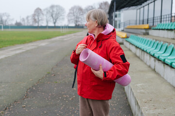Obraz premium An older woman holding a yoga mat stands on a sports field. She looks toward the empty seating area where people usually watch sports activities