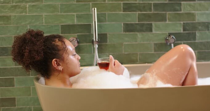 African American woman holding clear glass, shifting and reclining in tub with bubbles, relaxing
