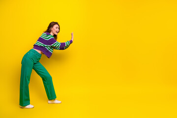 Young woman in striped pullover pushes against bright yellow background wearing green pants posing with energy for a fashion lifestyle image