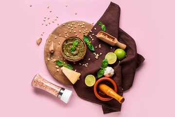 Wooden board with bowl of delicious pesto sauce, ingredients, mortar and scoop on pink background
