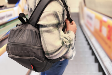 Close up of man with grey backpack standing on escalator. Person wearing plaid shirt in shopping mall or airport.
