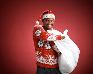 Smiling young Black man in a red Christmas sweater and Santa hat holding a large white shopping bag against a red studio background.