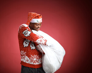 Smiling young Black man in a red Christmas sweater and Santa hat holding a large white shopping bag against a red studio background.