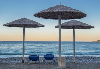 straw umbrella and sun loungers on a rocky beach at sunset in front of blue sea in Peloponnese Greece