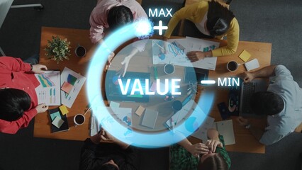A high-angle view of a diverse team engaged in a business meeting, analyzing graphs and discussing value metrics over a collaborative workspace filled with documents and devices. Raster