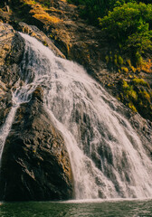 Close-up view of a powerful waterfall crashing down dark rocky cliffs. The dramatic flow of water highlights the natural texture and rugged terrain of a tropical landscape.