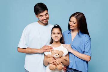 Young smiling lovely happy parents mom dad with child kid daughter girl wearing casual clothes hold hug plush toy teddy bear isolated on plain blue background. Family day parenthood childhood concept.