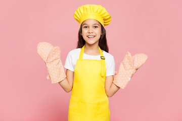 Small smiling happy surprised kid child girl housewife chef cook baker mother's helper wear yellow apron toque cap oven mitts look camera isolated on pastel plain pink background Cooking food concept