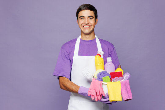 Young smiling happy man wear violet t-shirt hold basin with detergent bottles while doing housework tidy up look camera isolated on plain pastel light purple background studio. Housekeeping concept.