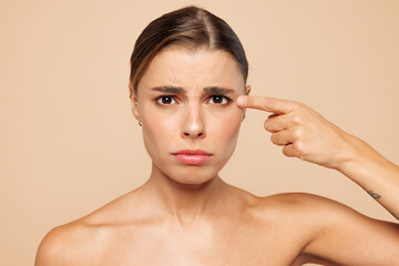 Naklejka premium Close up sad beautiful half naked topless young woman 20s with nude make up point index finger on isolated on beige color background studio portrait. Skin care healthcare cosmetic procedures concept.