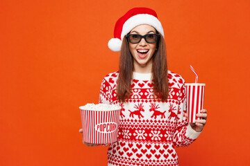 Merry young woman wear red Christmas sweater Santa hat 3d glasses watch movie film hold bucket of popcorn cup of soda pop in cinema posing isolated on plain orange background. Happy New Year concept.