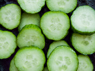Bunch of sliced fresh green cucumbers on the black background, top view of cucumber slices in the bowl covered with water drops.