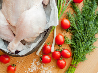 Whole raw chicken laying in the pan with parchment on the wooden table preparing for baking, surrounded by fresh vegetables.