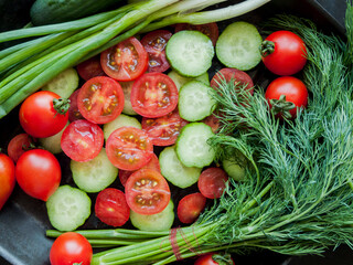 Fresh red cherry tomatoes, green leek, radish and cucumber in pan on black background, top view of vegetables covered with water drops.