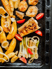 Oven pan with sliced baked potatoes, fish steaks and vegetables with onion on table.
