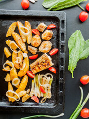 Oven pan with sliced baked potatoes, fish steaks and vegetables with onion on table.