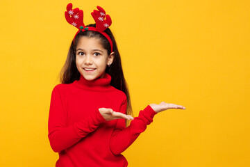 Merry little girl wears red clothes fun decorative deer horns on head posing point hands arms aside on area isolated on plain yellow background. Happy New Year Christmas celebration holiday concept.