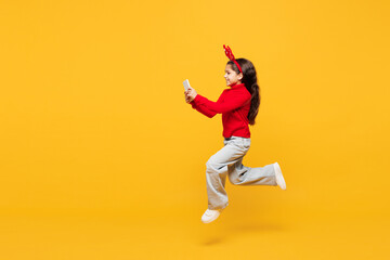 Full body merry little girl she wear red clothes fun decorative deer horns on head posing jump high...
