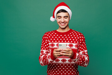 Merry young man he wear red warm Christmas sweater Santa Claus hat posing hold in hand use mobile...