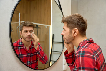 Man cleaning face with cotton pad while looking in mirror in bathroom. Concept of male skincare, facial cleansing, daily grooming routine and personal hygiene. © Lazy_Bear