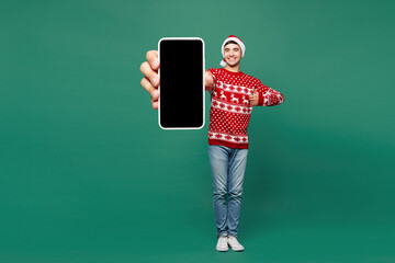 Full body merry young man wear red warm cozy Christmas sweater Santa Claus hat posing hold in hand use blank screen mobile cell phone isolated on plain green background Happy New Year holiday concept