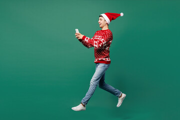 Full body merry young man wear red warm cozy Christmas sweater Santa Claus hat posing jump high hold in hand use mobile cell phone isolated on plain green background. Happy New Year holiday concept.