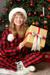 Pretty young woman in Santa hat with Christmas gift box sitting on floor at home