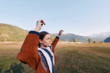 Fototapeta premium woman in nature with mountains and meadow background, autumn outdoor portrait wearing warm jacket, arms raised in joyful pose, serene landscape and soft daylight for travel lifestyle