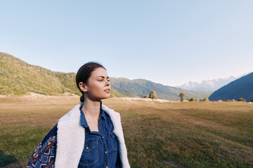 Fototapeta premium Woman in denim and shearling vest with backpack stands in a sunlit meadow near mountains, portrait with eyes closed enjoying nature, outdoor travel and calm landscape.