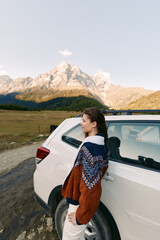 Naklejka premium Woman leaning against white car with cozy sweater in mountain landscape, outdoor travel and roadtrip portrait capturing scenic autumn adventure, peaceful nature and quiet exploration outdoors.
