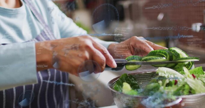 Senior woman picking up chef's knife, slicing cucumber into rounds and filling colander for cooking