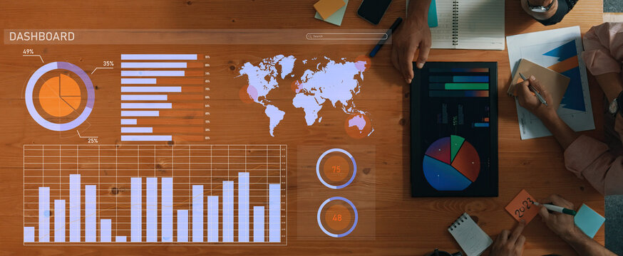 A top view of a business dashboard with various charts, graphs, and a world map on a wooden table. Hands are engaging with digital devices and notebooks. Axiom - Powered by Adobe