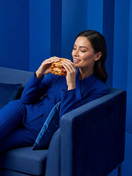 Woman in blue suit enjoying a sandwich on a blue couch, studio shot