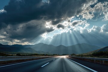 Asphalt road leads to light burst through dark, cloudy, mountain range backdrop