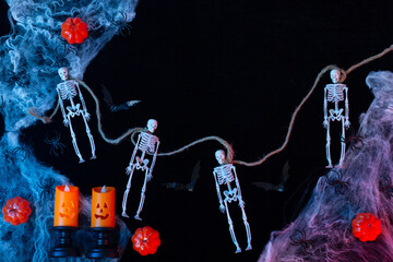 Halloween decoration: four skeletons hanging from a rope, on a black background, surrounded by cobwebs, pumpkins and candles, illuminated with purple and blue lights.